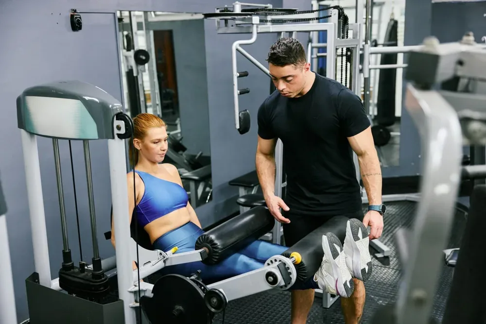 Une femme assise sur une machine de leg curl, jambes tendues et cuisses maintenues par un coussin, avec un homme debout à ses côtés.