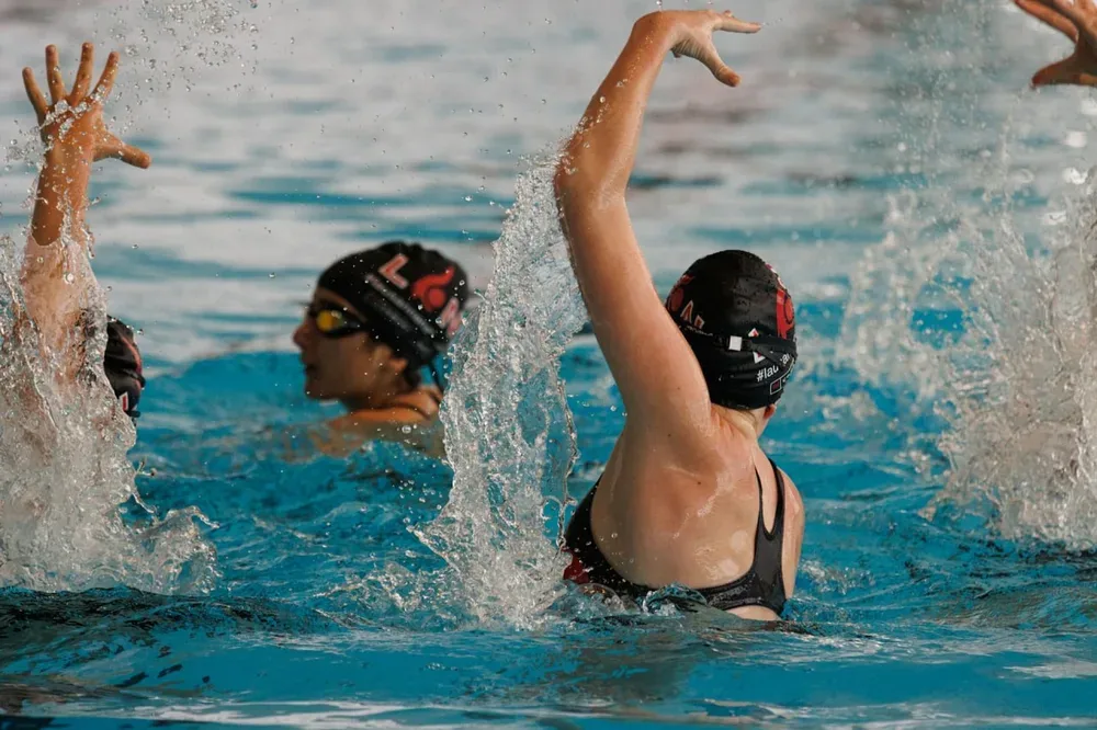 Sport en piscine : des nageuses en bonnet lèvent les bras dans un bassin, avec des éclaboussures autour d’elles.